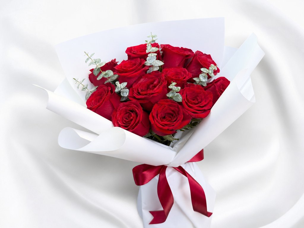 A dozen red roses with eucalyptus wrapped in crisp white paper tied with a red ribbon