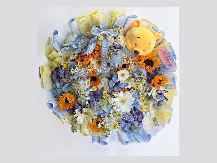 Round bouquet of sunflowers, daisies, and blue hydrangeas with butterfly accents and a honey-bear plush